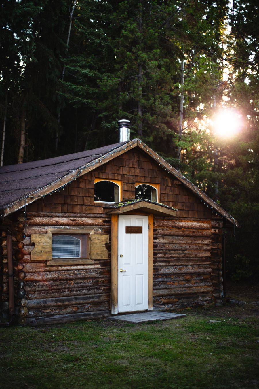 The Cottage, a log cabin with a white door, backlit by low sun through spruce