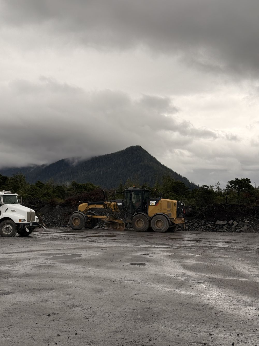 A motor grader and a truck on a wet gravel pad below a forested mountain