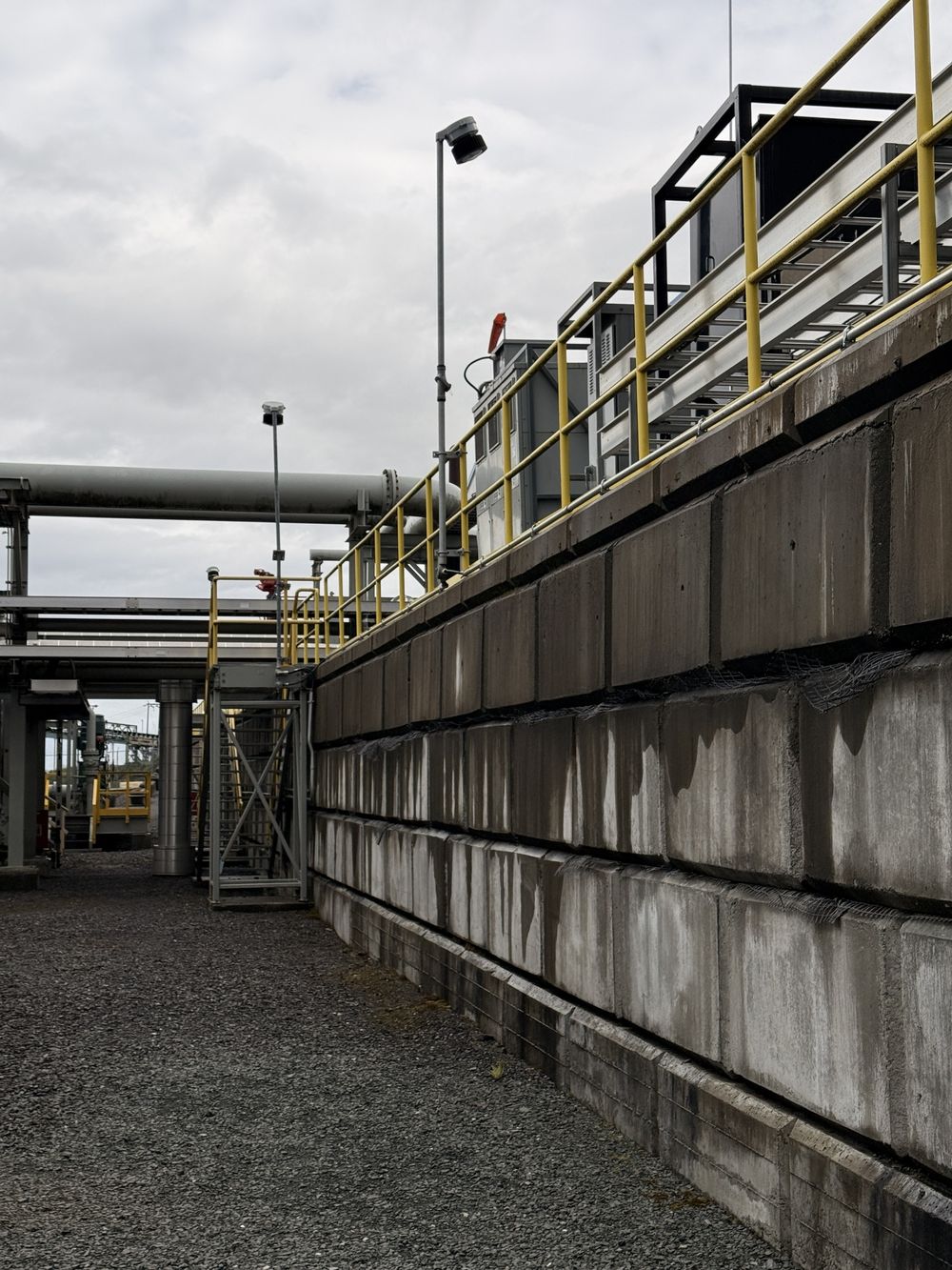 A block retaining wall with yellow handrails and pipe racks on an industrial site