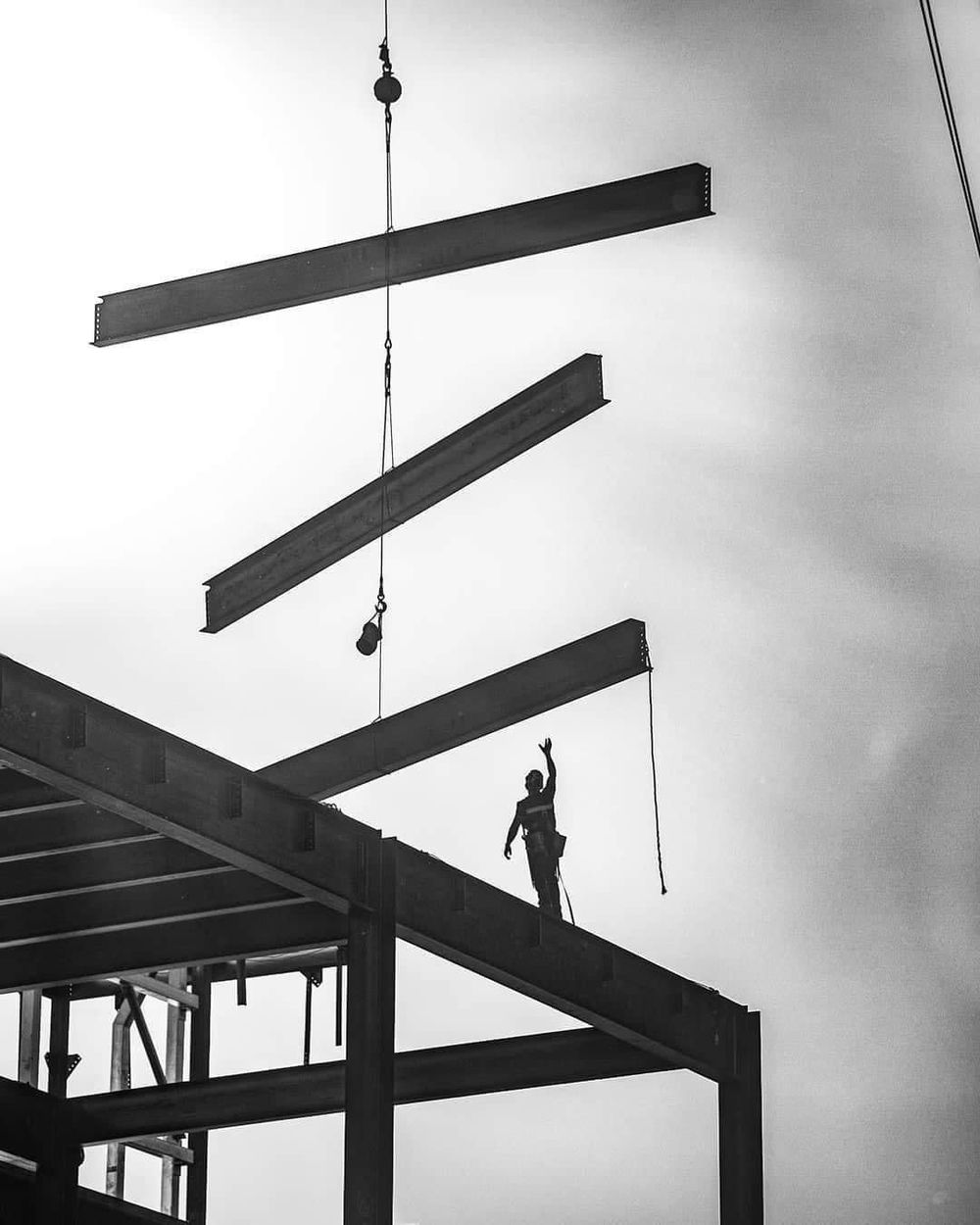 An ironworker on a steel frame signalling a crane as three beams are lowered into place